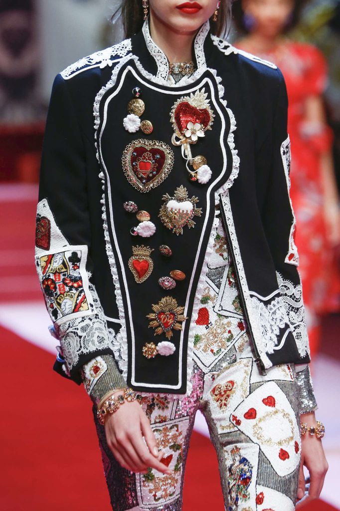 closeup shot of a model on a fashion runway wearing a richly embellished dolce & gabbana jacket and pants. the jacket is black with intricate lace detailing and features numerous ornate appliqués in the shape of hearts, playing cards, and other decorative elements. the pants are similarly decorated with playing cards and sequins.  the model is also wearing several bracelets and earrings. the background is a blurry red carpet, suggesting a fashion show setting. the overall look is opulent and luxurious, showcasing highfashion craftsmanship.