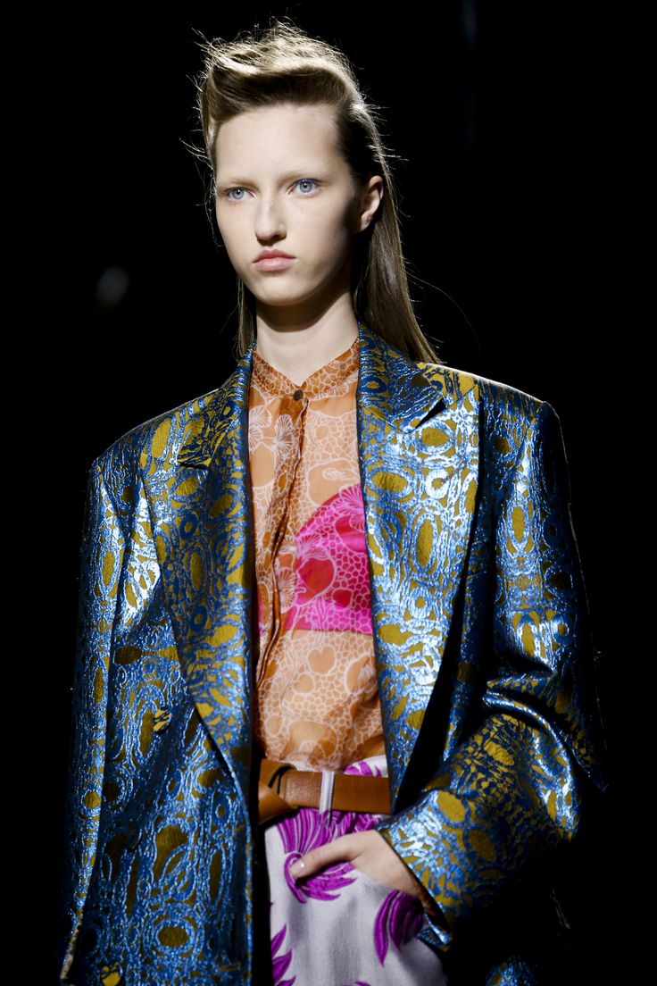 the image showcases a fashion model on a runway, wearing a striking ensemble. the centerpiece is a long, oversized blazer in a rich teal and gold brocade fabric with a distinct textured, almost floral pattern.  underneath, she wears a sheer blouse in shades of orange and pink with a discernible pattern. the blouse features a bright pink section that resembles a bra.  a brown leather belt cinches her waist.  her pants are partially visible, showing a white and purple floral print. the model's hair is styled in a sleek updo, and her makeup is clean and understated, emphasizing her features. the overall aesthetic is one of high fashion, sophistication, and bold color choices. the background is dark and blurred, focusing attention entirely on the model and her outfit.