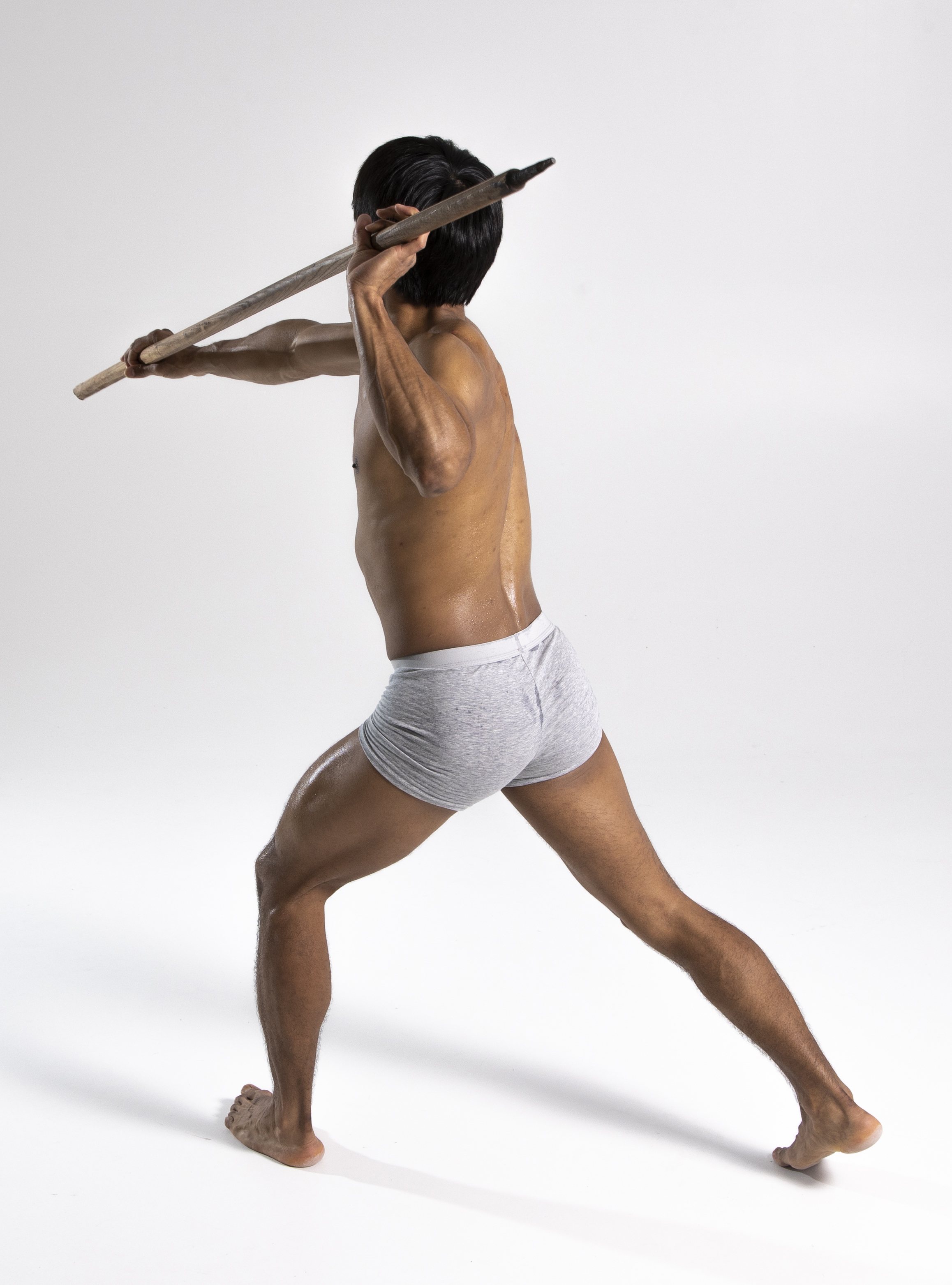 a lightskinned, athletic man in grey shorts stands indoors against a white background, holding a spear and getting ready to throw it. the man is barechested and barefoot. the shot is a full shot taken in a studio.