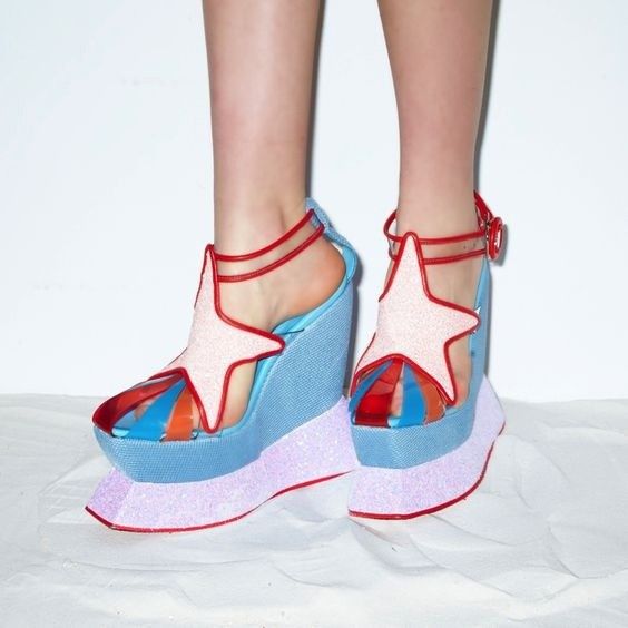 a closeup shot shows a pair of highfashion wedge heels with a whimsical and playful design. the shoes feature a light blue wedge heel, a pink glitter platform sole, and a red starshaped design on the front. clear plastic accents reveal a colorful striped pattern beneath the star. red straps secure the shoes around the ankles. the shoes are worn by someone standing on a white surface, and the background is plain white.