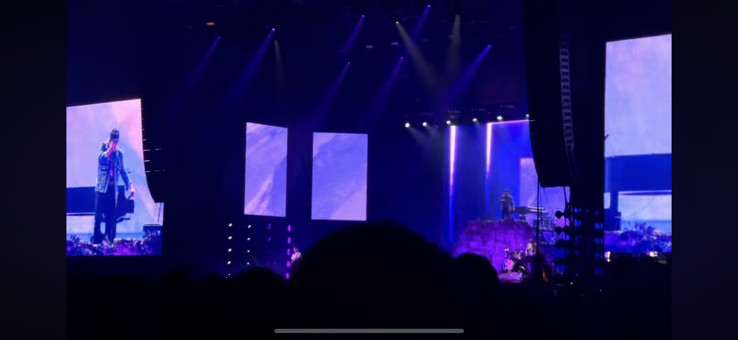 a concert stage with a singer standing in front of a large screen, lit by purple spotlights. the audience is silhouetted in the foreground, with the sound system on the right side of the image.