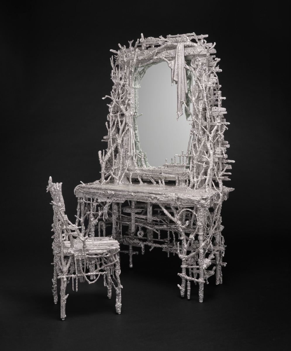 the image shows a metal sculpture of a dressing table with a mirror and a chair. the entire set is silver in color and appears to be handcrafted, featuring an intricate design that mimics twigs and branches. the mirror is framed by a similar structure, and a draped cloth is partially visible within the reflection. the background is black, which highlights the metal's texture.