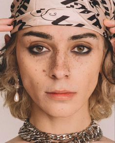 a young woman with blonde hair and freckles poses for the camera in a studio. she is wearing a silver chain necklace and a silver nose ring. she has  dark eye makeup and her hair is styled in a headband. her facial expression is serious and she is looking directly at the camera.
