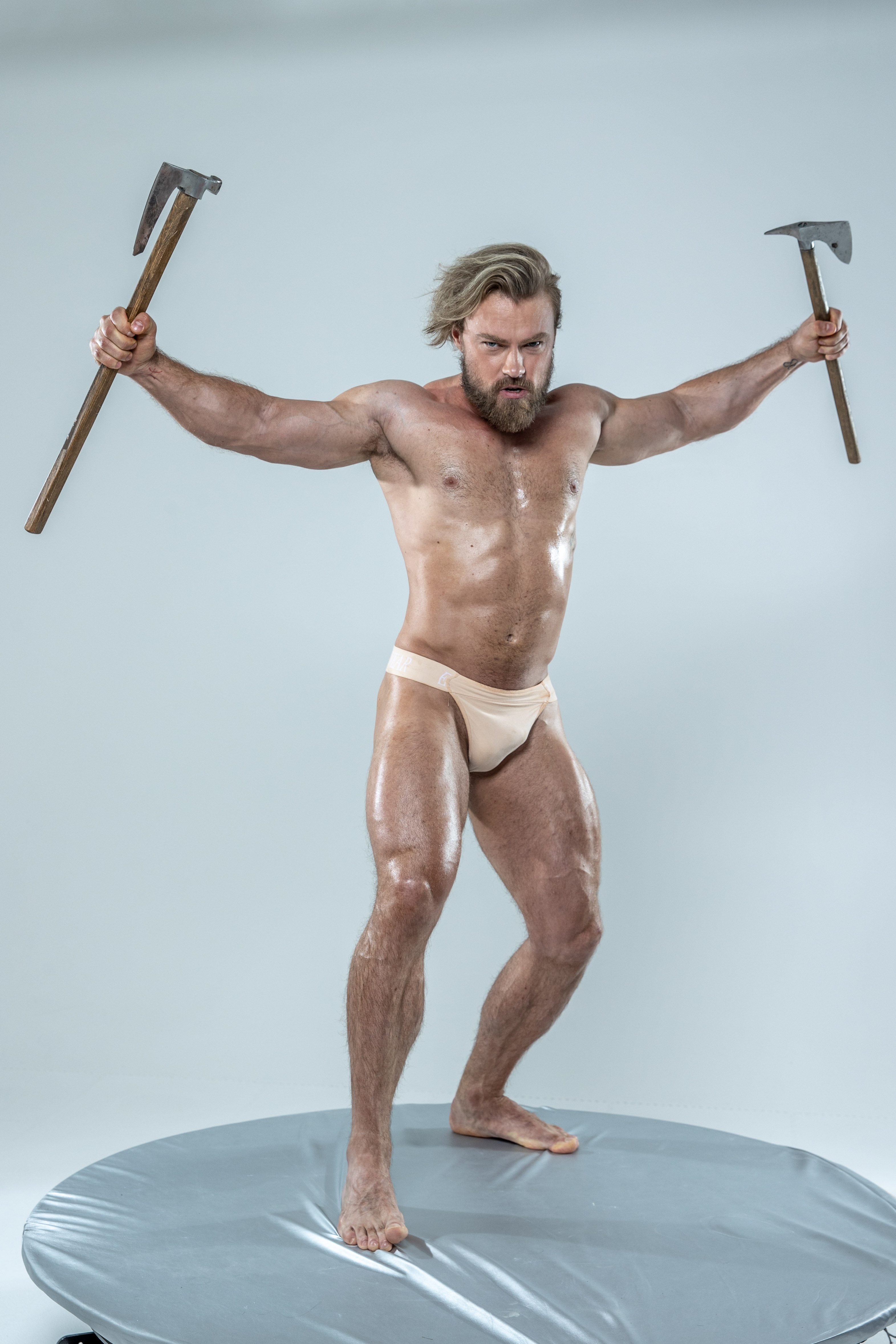 a muscular, bearded man with long blond hair stands on a round grey platform against a light grey background.  he wears only a light beige loincloth and brandishes two axes  one in each hand  held high. his body is glistening, suggesting he is oiled or sweating. the overall impression is of a fierce warrior, possibly a viking or barbarian, ready for battle.