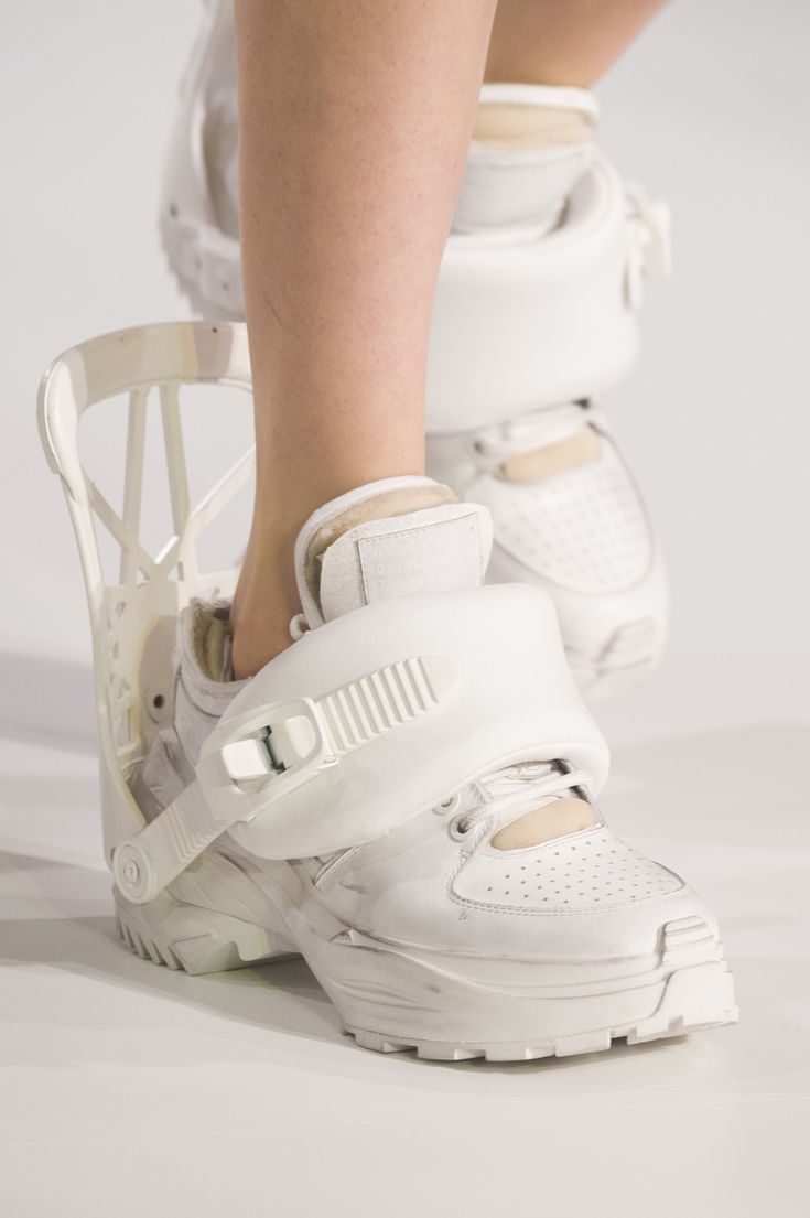 a close up shot of a person's leg wearing white sneakers. the sneakers are bulky and appear to be made of white leather. the person is wearing a white outfit, and their skin is white. the sneakers have a unique design with a large, chunky sole and a strap that goes around the ankle. the person is standing on a white surface.