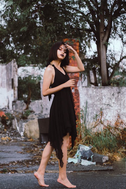 a woman stands barefoot in front of a dilapidated building, wearing a sleeveless black dress. she has dark lipstick and a bob haircut. her hand is on her forehead as if shielding her eyes. the background features ruins, a tree, and an urban grunge setting.