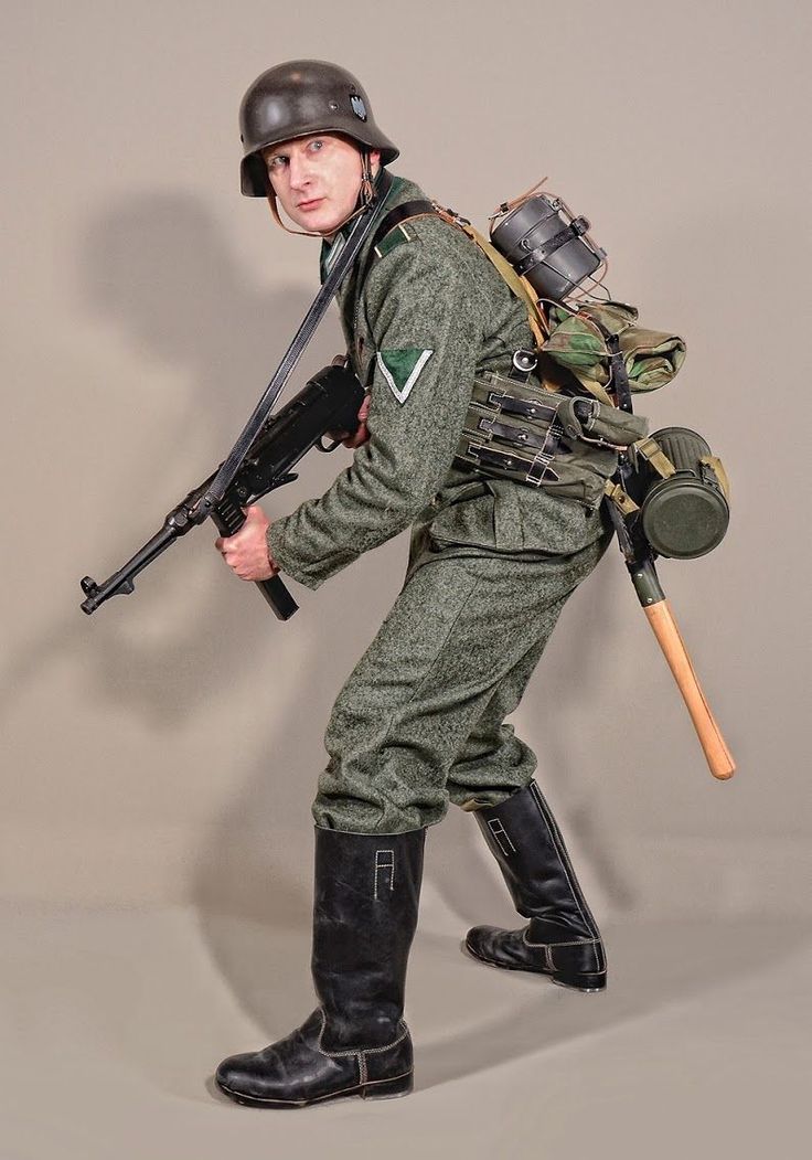 a man dressed in a german wwii era soldier uniform is posing holding a rifle. he is wearing a helmet, boots, and a backpack with gear, all of which are consistent with the time period. he is in a studio setting with a neutral background.