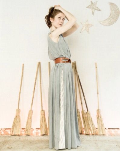 a woman is standing in front of a white wall decorated with a star and moon. she is wearing a long, light blue dress with a brown belt, and her hair is pulled back in a ponytail. several brooms are leaning against the wall behind her. she is holding her hands near her head.