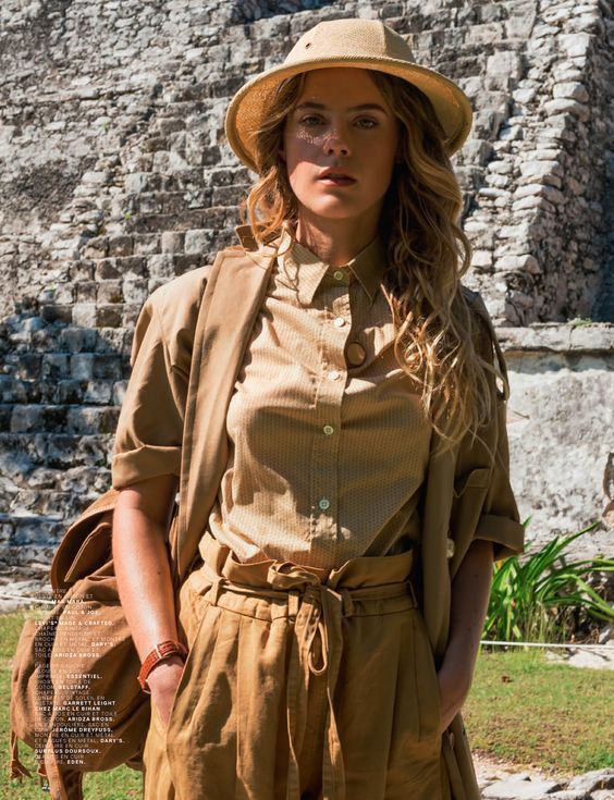 a fashion photograph of a young woman, possibly a model, in a stylish khakicolored outfit. she is wearing a safaristyle hat, a buttondown shirt, a light jacket, and loose, highwaisted pants. a backpack is slung over one shoulder.  she is standing in front of ancient stone ruins, suggesting a travel or adventure theme.  the overall style is relaxed yet chic. the setting appears to be outdoors in bright sunlight.