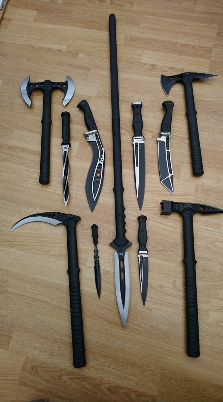a variety of modern tactical weapons are laid out on a light brown wooden surface. there is a double bladed axe, a tomahawk, a karambit, a sword, a warhammer, and a war scythe.
