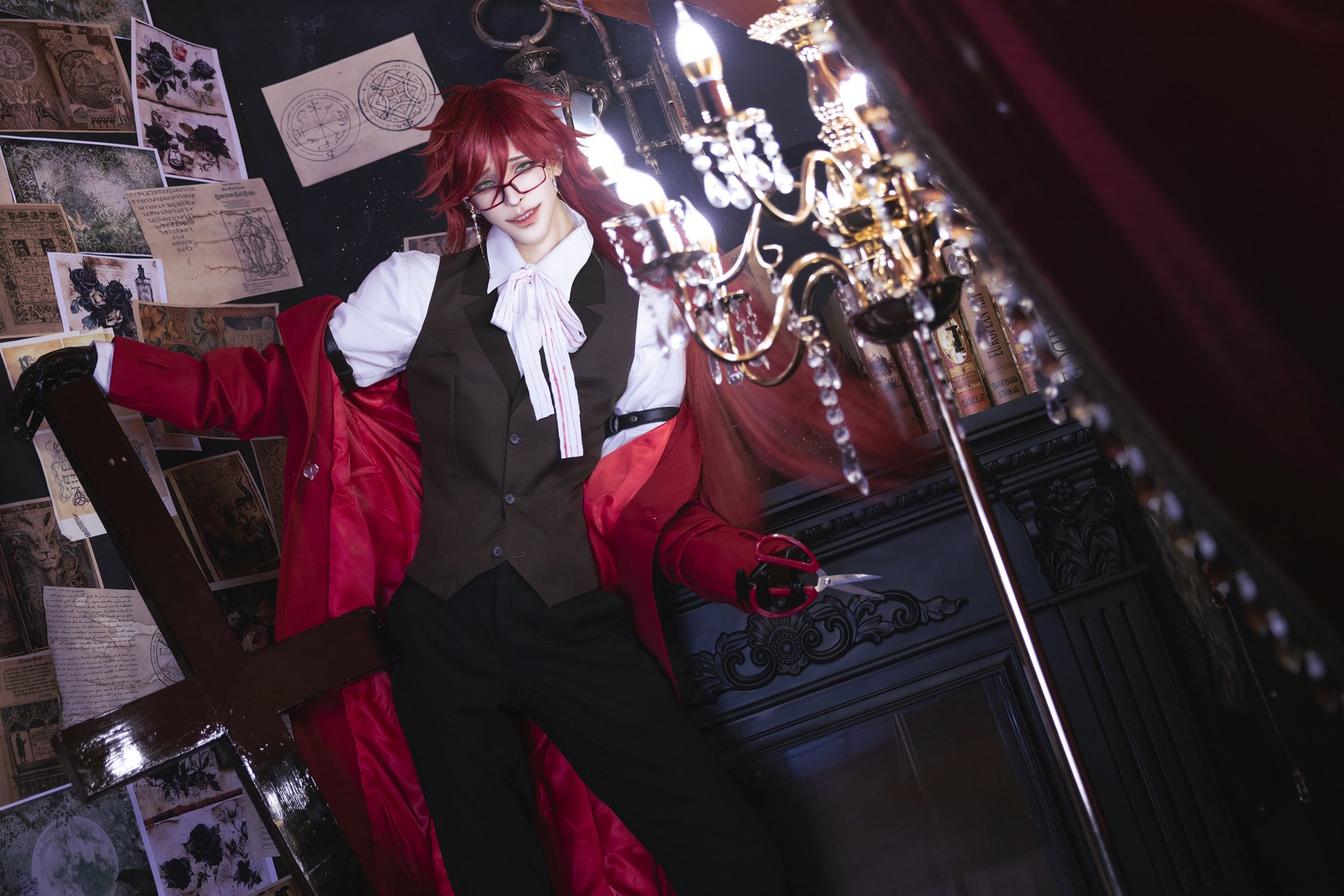 a cosplayer dressed as ciel phantomhive from the anime/manga black butler is standing in a dark and gothic setting. they are wearing a black suit with a red vest, a white shirt, and a black bow tie. they are also wearing red gloves and a red cape. they are holding a cross and scissors, and they are looking directly at the camera.
