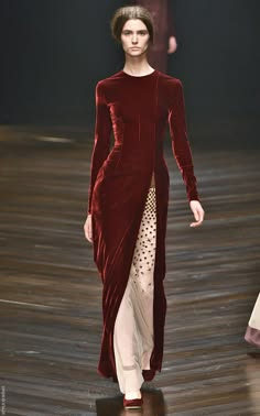 a woman in a long, red velvet dress poses on the runway. she is wearing high heels and has her hair styled in a simple, elegant way. she is looking down at the camera.