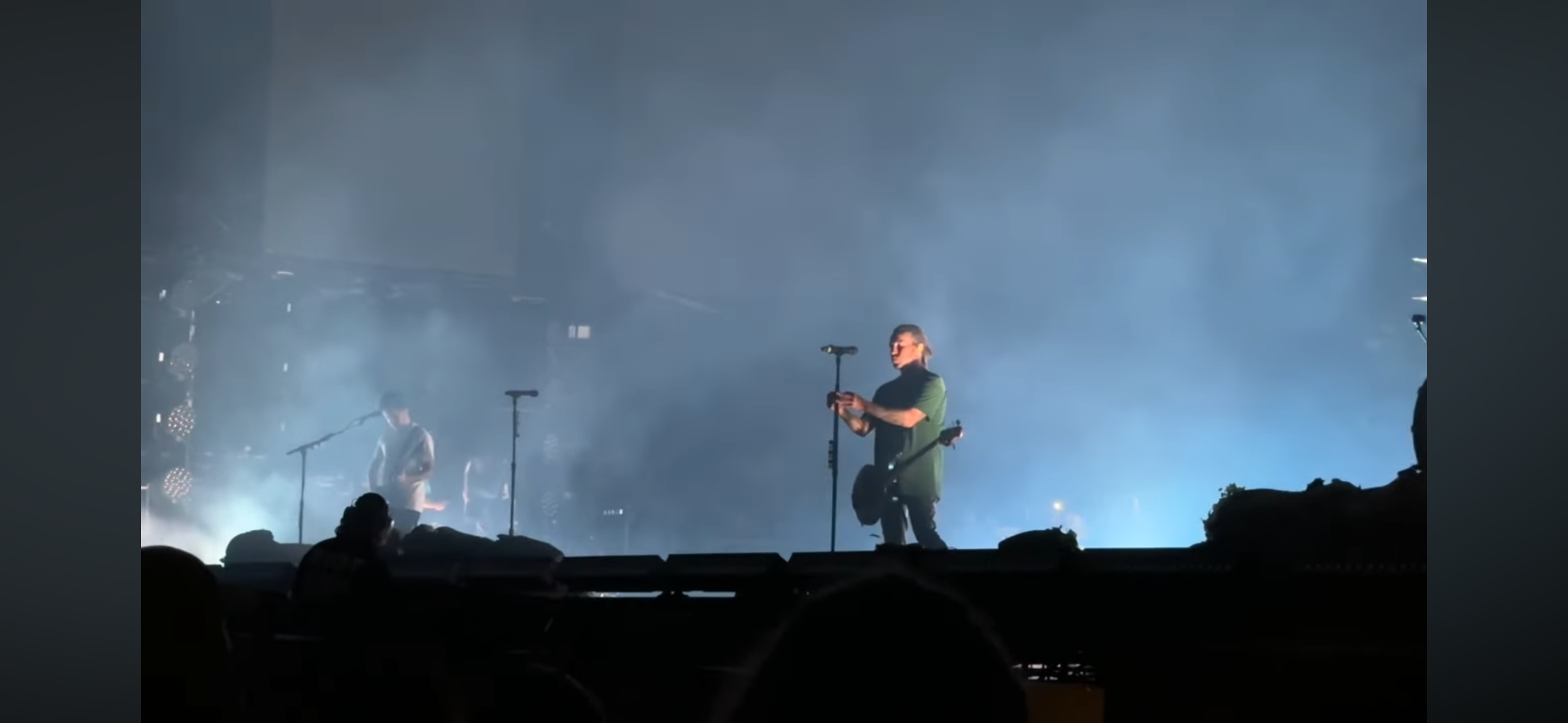 a concert scene showing the band on stage with the lead vocalist singing into a microphone and holding a guitar. the stage is lit with fog and lights, and the audience is silhouetted in the background. the stage is a large stage with a microphone stand in the center, and a microphone stand on the right. the band has 2 people in the center of the stage, one on the left with a guitar, and the other one in the center with a microphone. there are a few rows of lights on the upper edge of the stage. the venue is very dark, with the lights from the stage providing a contrast. the audience is very dark, with the only thing that stands out are the heads of the people in the front row.