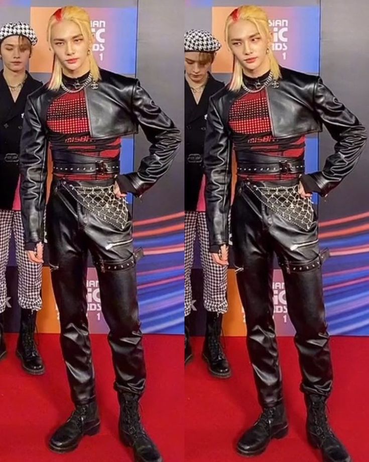 the image shows a man with blonde hair and a red stripe in his hair, posing on a red carpet. he is wearing a black leather jacket, a mesh top, a chain belt, and black leather pants with boots. another man wearing a checkered hat is visible in the background. the scene appears to be from the asian music awards.