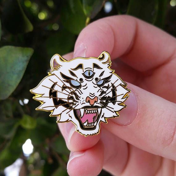 a hand holds a white enamel pin in the shape of a roaring white tiger's head with three eyes. the pin has gold edges, with black stripes, blue eyes, a pink tongue, and sharp teeth. the hand holding the pin has pink fingers, and the background features blurred green leaves.
