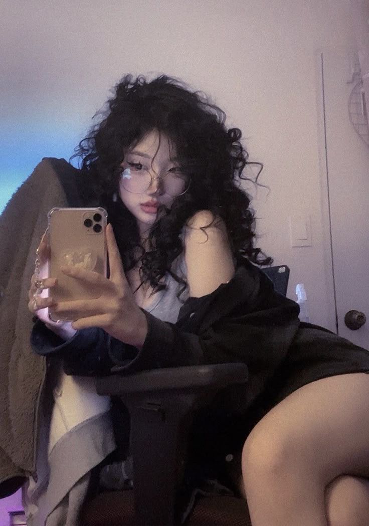 a woman with curly hair and glasses takes a selfie indoors, sitting in a chair while wearing a stylish outfit and makeup. she is holding her phone and wearing rings.