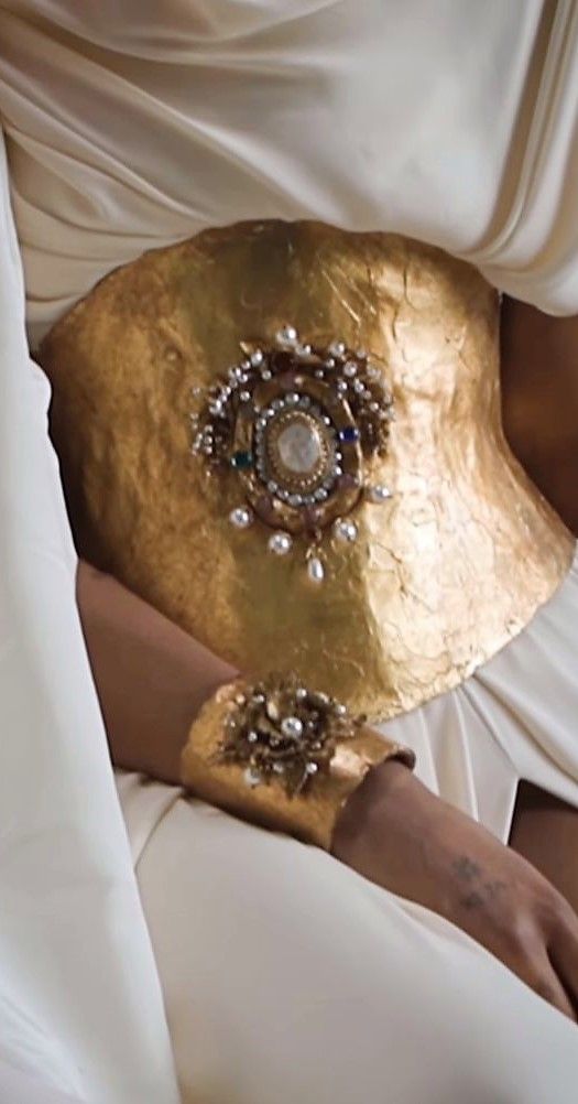 a closeup of a woman's arm wearing a gold bracelet and a gold belt with a large ornate jeweled centerpiece. the woman is sitting in a white dress with her arm resting on her lap. the lighting is soft and smooth, highlighting the details of the jewelry and the woman's skin.