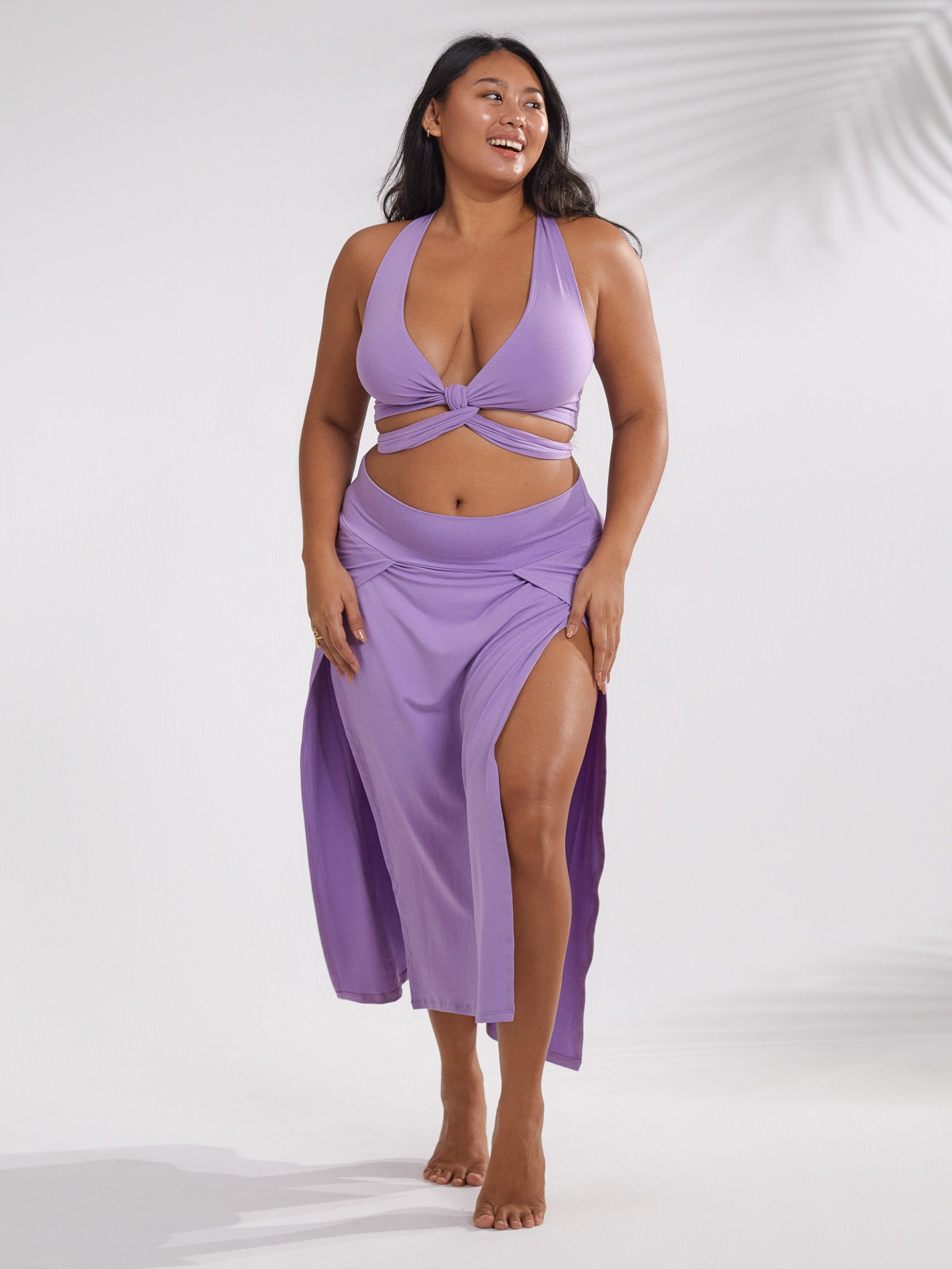 a fullbody shot of a plussize woman confidently posing in a stylish purple swimsuit and skirt set.  the swimsuit features a halter top with a tie knot at the center, and the skirt is a maxilength with a high waist and side slits. she is smiling and looks directly at the camera against a simple white backdrop with subtle shadowing. the overall impression is of summer beachwear and body positivity.