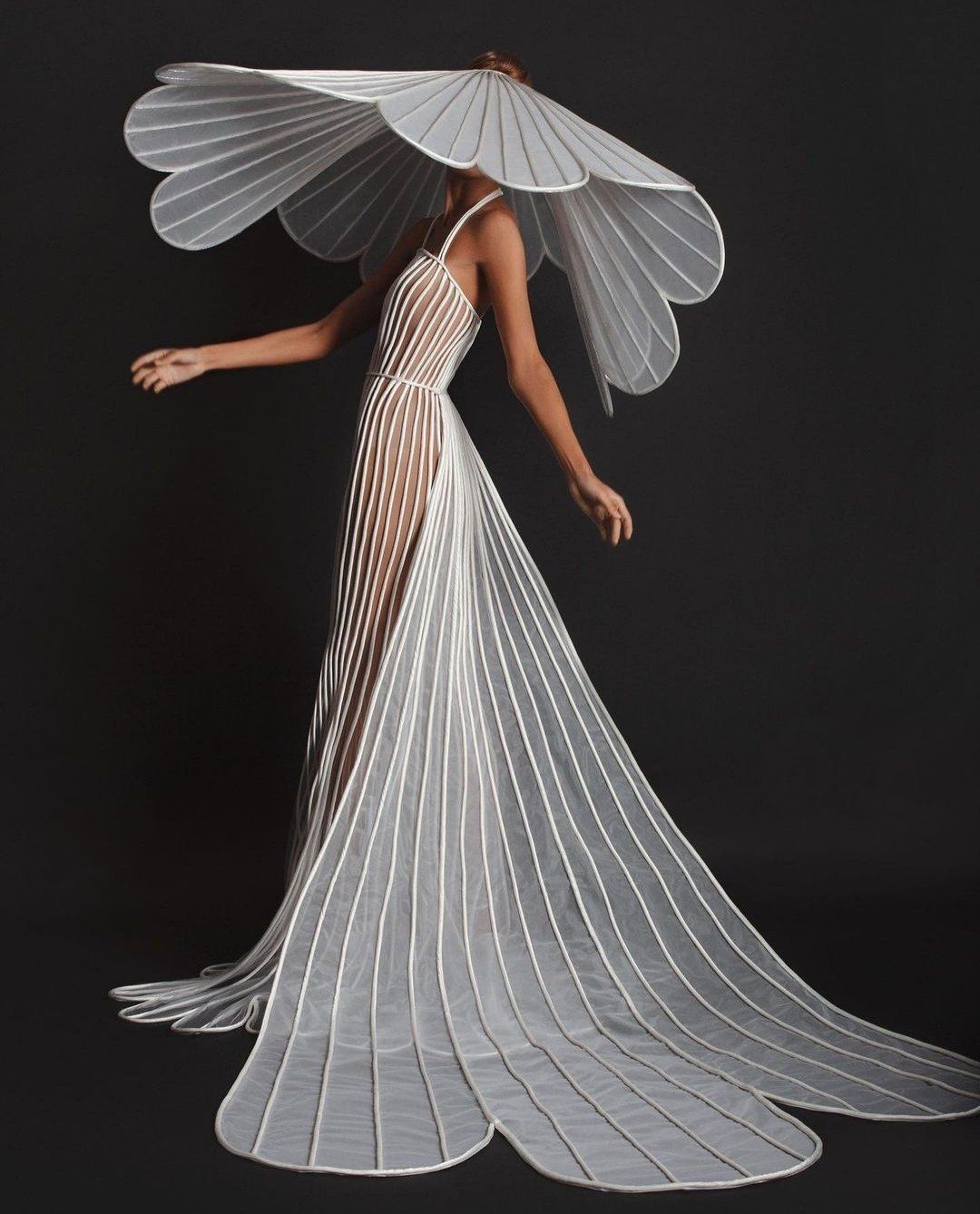 a woman is modeling a white, avantgarde gown with a large, flowerlike structure covering her head. the dress is made of sheer fabric with vertical white lines creating a minimalist and elegant look. the gown extends into a long train with petalshaped edges. the background is dark, emphasizing the dress and the model.