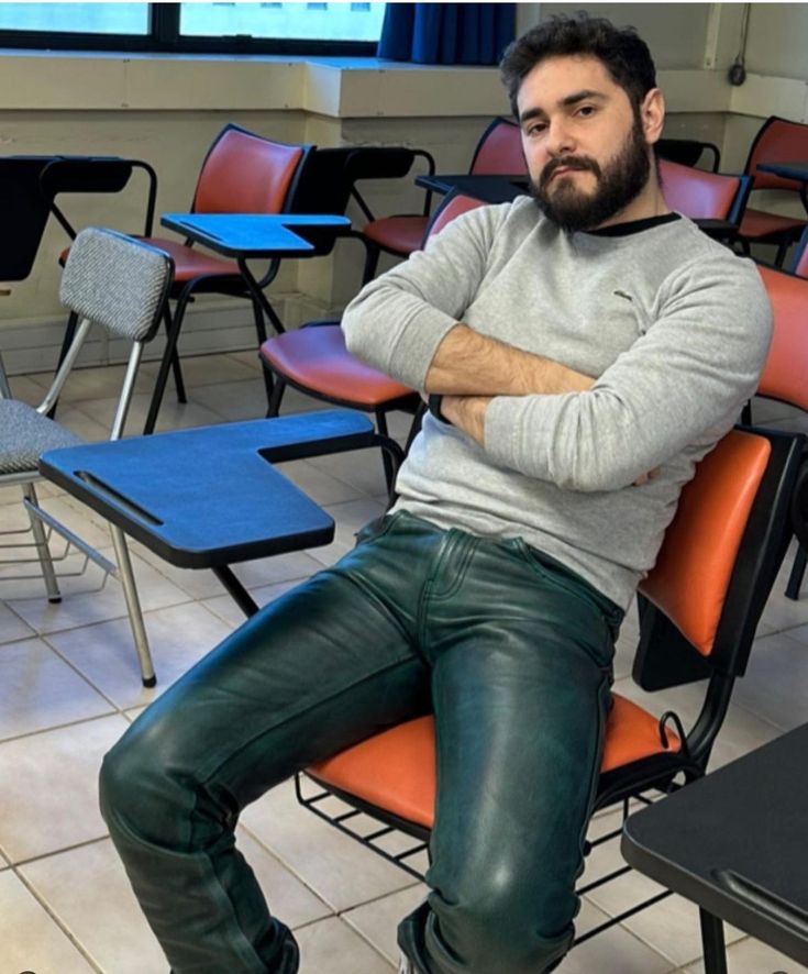 a man with a beard, wearing a grey sweater and dark green leather pants, sits with his arms crossed in a classroom. he is seated in an orange chair at a desk, surrounded by other empty desks and chairs. the setting appears to be a college or university classroom.  the man's expression is serious.