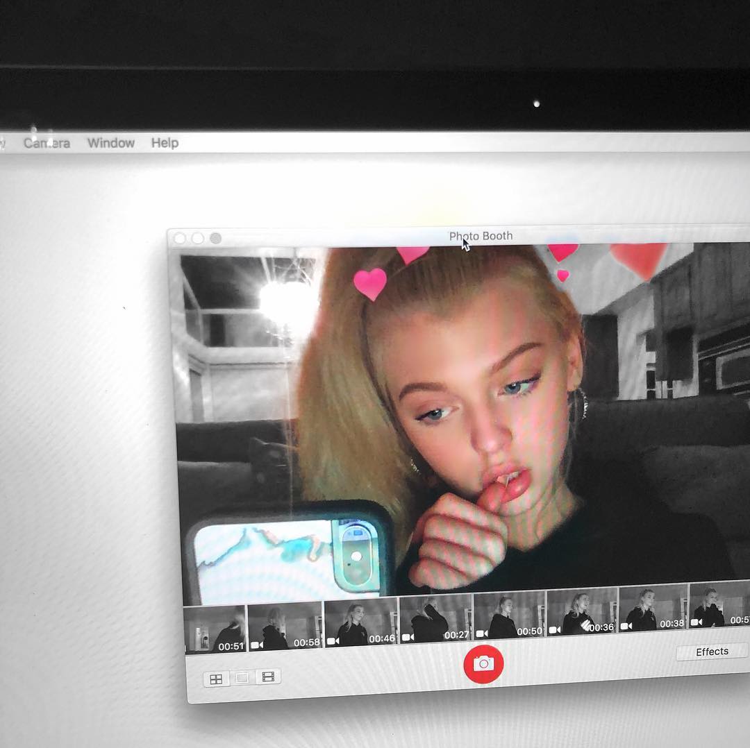 a computer screen shows the macos photo booth application with a young woman using a digital filter that adds pink hearts above her head. she is holding a smartphone in the foreground, and several recorded clips appear at the bottom of the application window. the computer's menu bar with options like "camera," "window," and "help" is visible at the top.