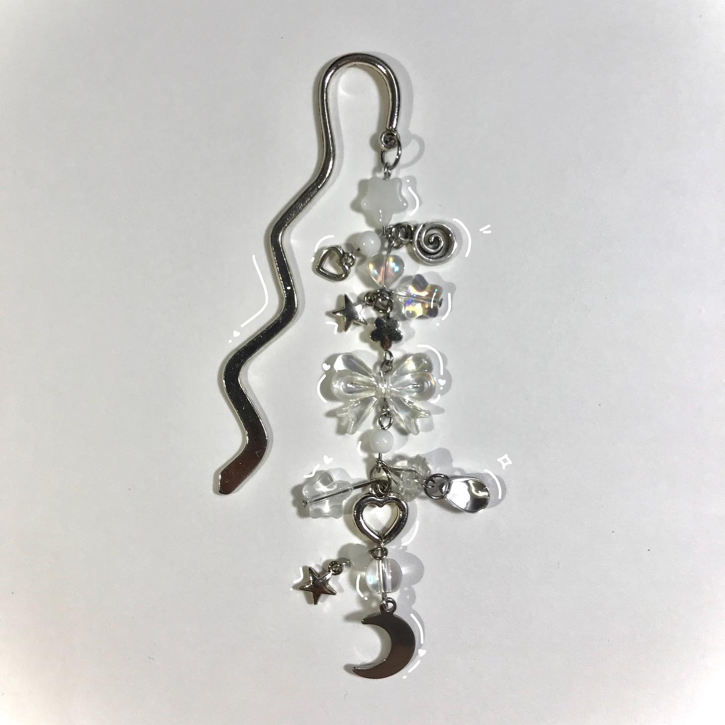 a silver bookmark with a wavy design is adorned with various charms, including a moon, star, heart, and bow, along with clear and iridescent beads. the charms are attached to the bottom of the bookmark, creating a dangling, whimsical effect. the aesthetic is sparkly and celestial.