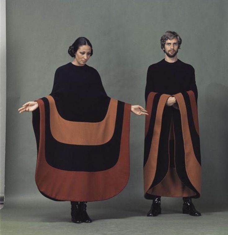 a vintage fashion photograph features a man and a woman, both dressed in black tops and matching capes with brown and black geometric designs. the woman is on the left with her arms outstretched, showcasing the full width of her cape. the man is on the right with his hands folded, holding the edges of his cape together. they both wear black boots and stand against a plain gray background. the image evokes a retro style from the 1970s.