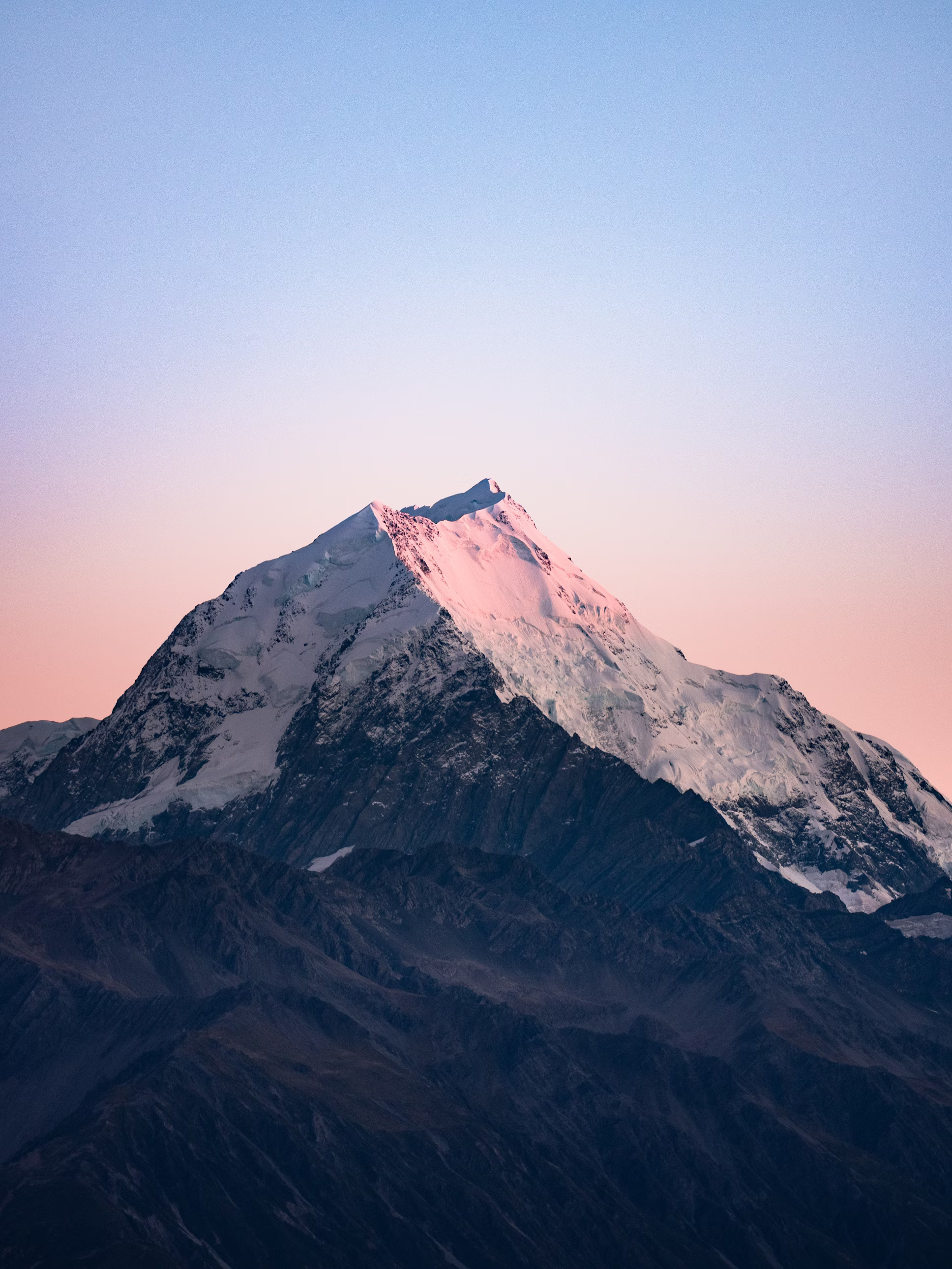 a majestic, snowcapped mountain dominates the frame, set against a serene twilight sky. the setting sun casts a warm, pinkish glow on the peak, while the lower slopes fade into cool, bluetinged shadows. the foreground features the dark silhouettes of other mountains, emphasizing the vastness and grandeur of the scene.