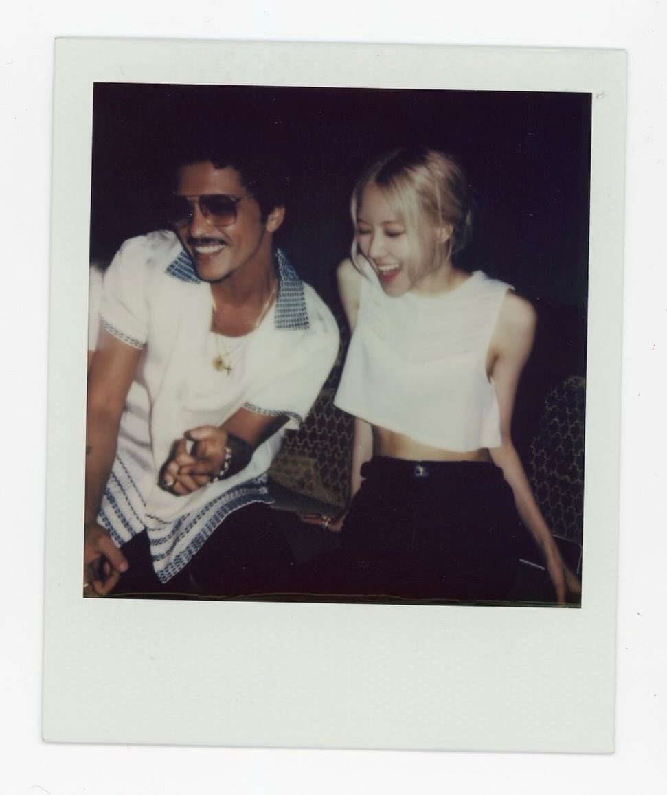 the image is a polaroidstyle photograph showing a closeup of a man and woman, identified as bad bunny and rose from the kpop group blackpink, sitting close together. they are both smiling and laughing. the man is wearing a white shirt with a collar, sunglasses, and a necklace. the woman is wearing a white crop top and black pants. the background is dark and indistinct. the overall mood of the photo is casual, affectionate, and joyful. the polaroid style adds a vintage and nostalgic feel to the image.