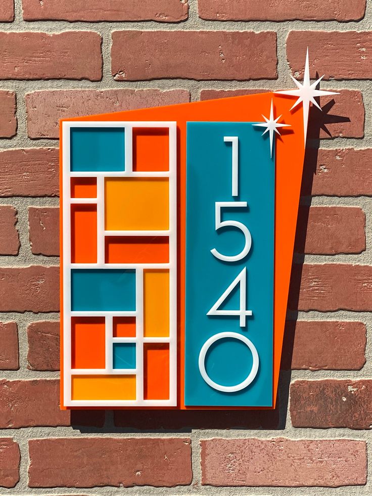 a modern house number sign with a geometric design. the sign is made up of orange, teal, and white squares and rectangles and features the number 1540 in white against a teal background. the sign is mounted on a brick wall.