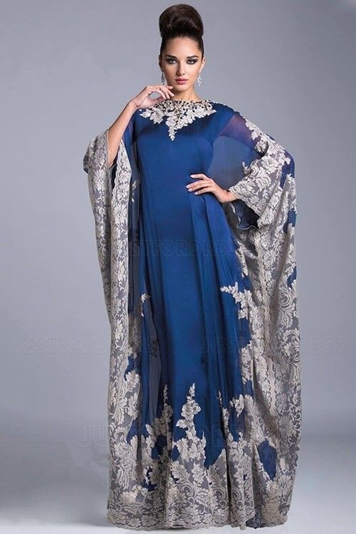 a full shot of a woman modeling a stunning blue maxi dress. the dress features a high neck, long sleeves, and intricate lace detailing along the edges. the lace is a light beige color which contrasts with the deep blue of the dress. the woman has her hair styled up in a bun and is wearing minimal jewelry. the background is a solid light grey.  the overall style suggests it would be appropriate for formal occasions or special events.