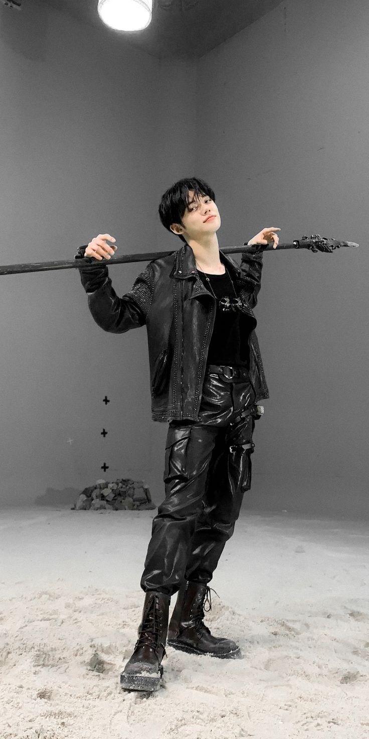 a young man in black leather jacket and pants poses in front of a white background, holding a long black stick with both hands. he is wearing black leather boots and has his hair styled in a short, messy cut. the photo has a cool and edgy feel.