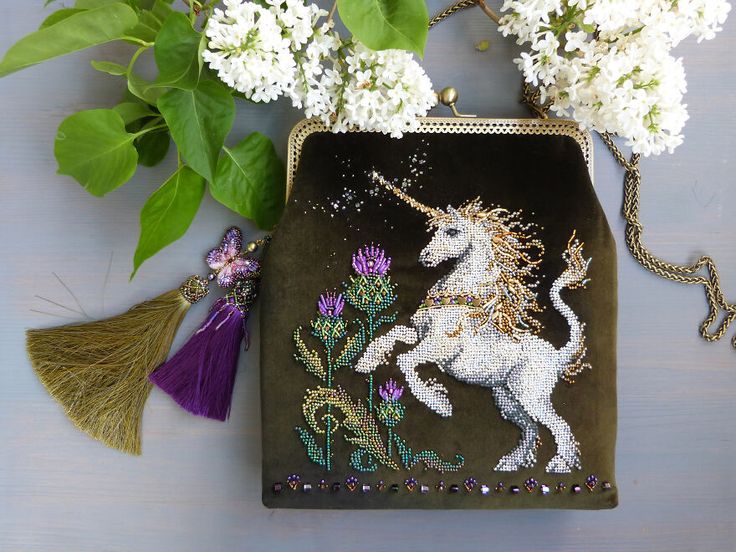 a closeup shot of a green velvet purse with a beaded unicorn embroidery design. the purse is decorated with a golden chain and two tassel charms. the purse is sitting on a gray surface with a branch of white flowers to the top left of the purse.