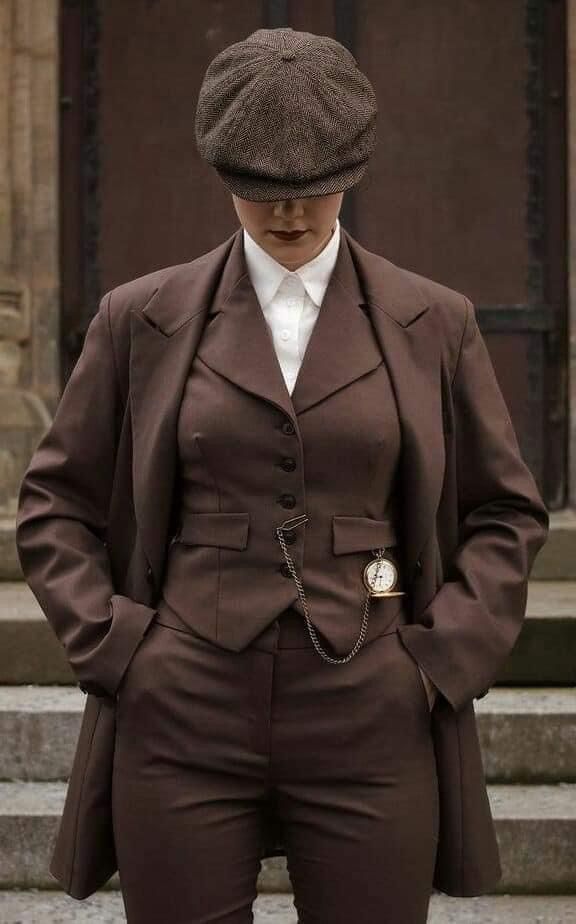 a woman in a vintage brown suit poses for the camera. the suit is a threepiece, with a jacket, vest, and pants, and she is wearing a brown cap. she has a serious expression on her face, and she is looking at the camera. the suit is welltailored and fits her perfectly. she is a fashionable and elegant woman who knows how to make a statement. she has a pocket watch chain hanging from her vest, and she is looking like a very fashionable vintage lady.