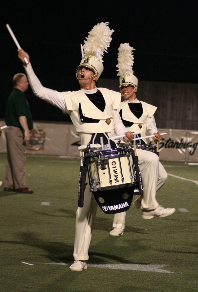 two young drum majors in white and black uniforms march in a drum line during a football game. they are holding drumsticks and smiling as they march.