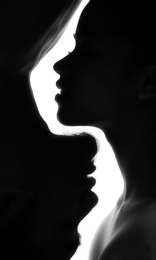 the image is a dramatic black and white silhouette of a couple intimately close to each other. their profiles are visible, their faces almost touching, suggesting a moment of intense connection and romance.  the strong contrast between the dark silhouettes and the bright background emphasizes the closeness and intimacy of the scene.  the lack of detail in the silhouettes adds to the mysterious and evocative nature of the photograph.