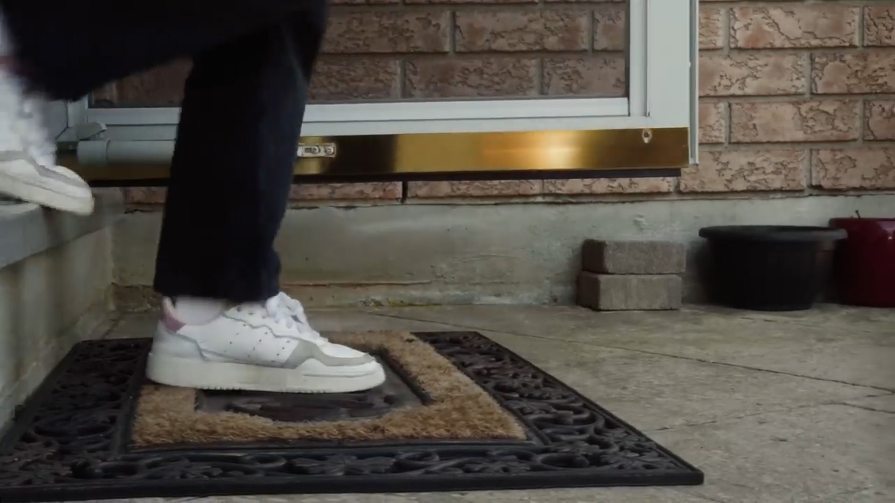a person wearing white adidas sneakers is stepping onto a doormat in front of a house. the image is taken from a low angle, showing the person's legs and feet. the lighting is natural.