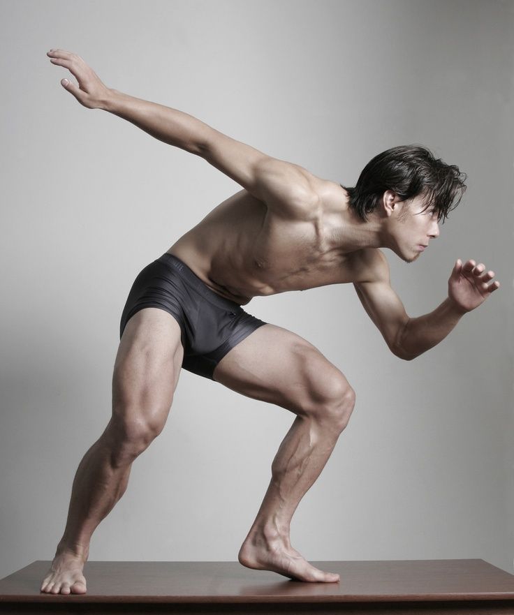 a muscular man in black underwear is posing with one leg extended, his body is contorted with his arms extended and hands curled. he is standing on a wooden platform with a grey background.