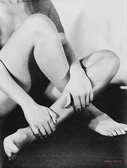 a black and white photograph depicting a nude female figure seated, her legs crossed and hands gently touching her lower legs. the image is intimate and focuses on the texture and form of the woman's body. the lighting and shadows create a sensual mood, highlighting the curves and contours of her skin. the overall style is reminiscent of classic fine art photography.
