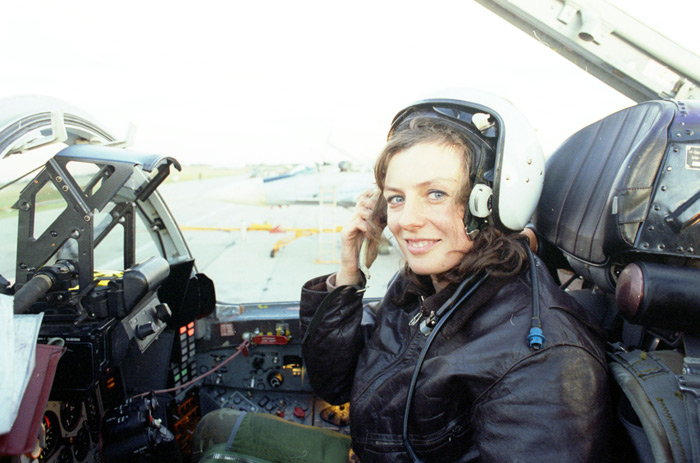 a woman pilot sits in the cockpit of a fighter jet wearing a headset and helmet, smiling and looking at the camera. she is wearing a dark leather jacket. the cockpit is filled with various controls and instruments. the image is likely taken during an air show or other flight event.