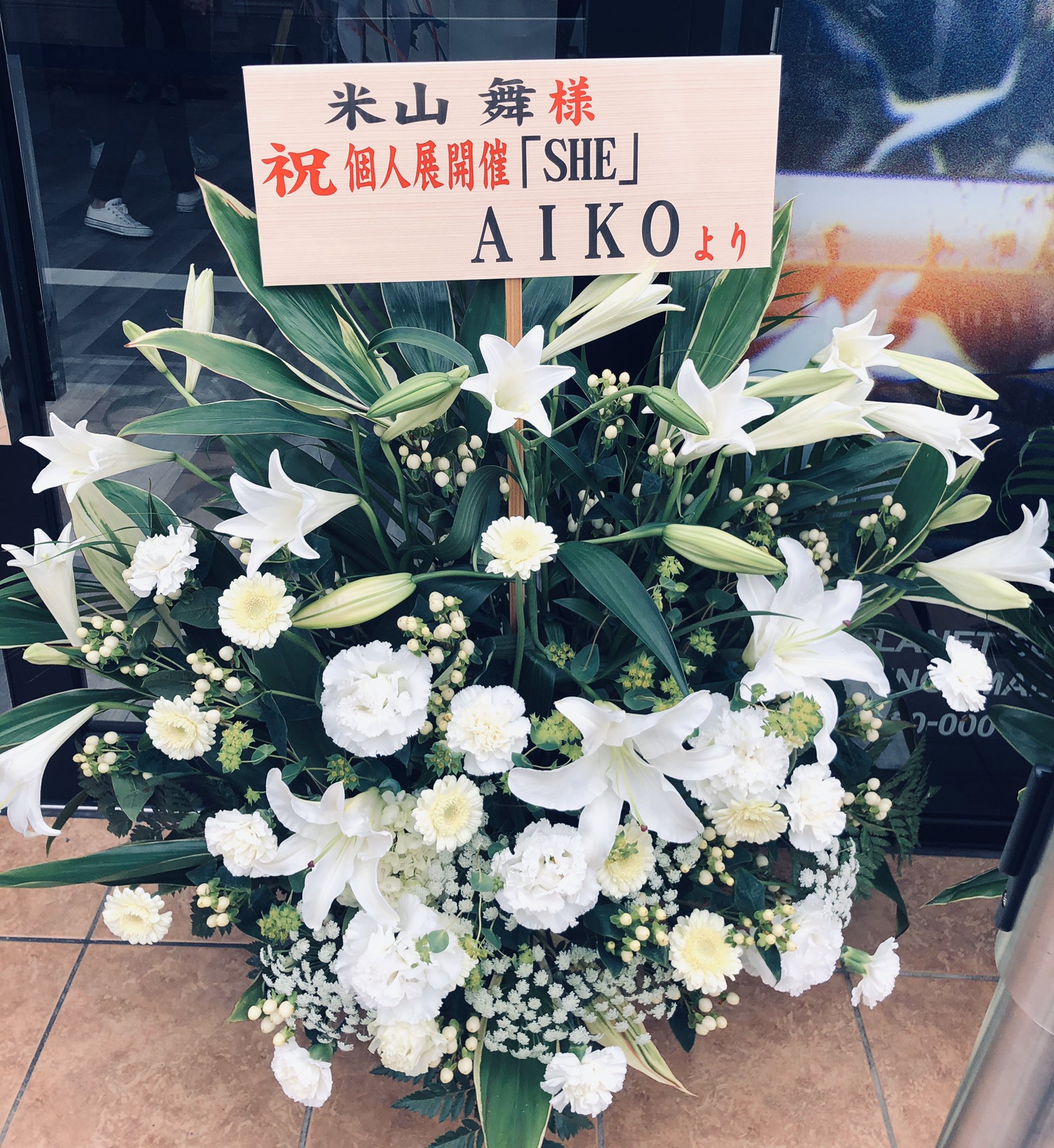 a large bouquet of white flowers in a floral foam base sits on a tiled floor. the flowers are primarily lilies, with some carnations and greenery mixed in. there is a sign in the arrangement that says "[illegible japanese] 米山 舞樣 祝個人展開催「she」 aikoより", which translates to "[illegible japanese] ms. mai yoneyama congratulations on your solo exhibition "she" from aiko". the background is blurry and out of focus, but appears to be the interior of a building.