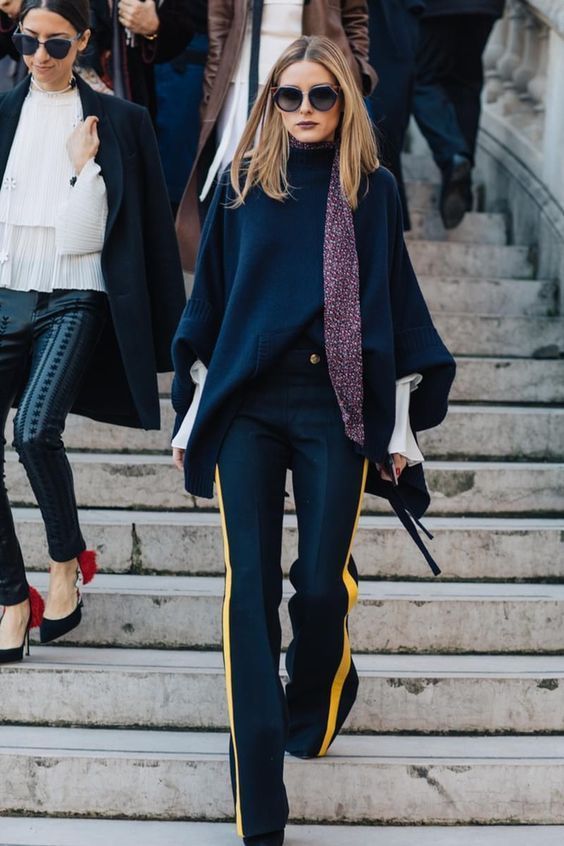 a stylish woman wearing a navy blue poncho, dark blue pants with yellow stripes down the sides, a purple scarf, and sunglasses is walking down a set of stairs.  another woman in a white top, black leather pants, and a dark coat walks behind her. the background suggests a parisian street scene. the overall impression is one of sophisticated and trendy street style.