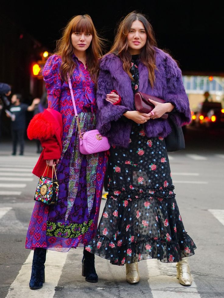 wo women are standing on a city street. one woman is wearing a purple fur coat over a black floral maxi dress with gold boots. the other woman is wearing a purple and pink floral maxi dress with velvet boots. both women are carrying bags. the image is a street style photo.