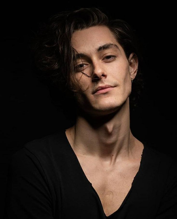 a portrait of a man with dark hair and a black shirt against a dark background. he has a confident expression and a welldefined jawline. his hair is styled in a slightly messy way, and he has an earring in his left ear. the focus is on his face and upper torso.