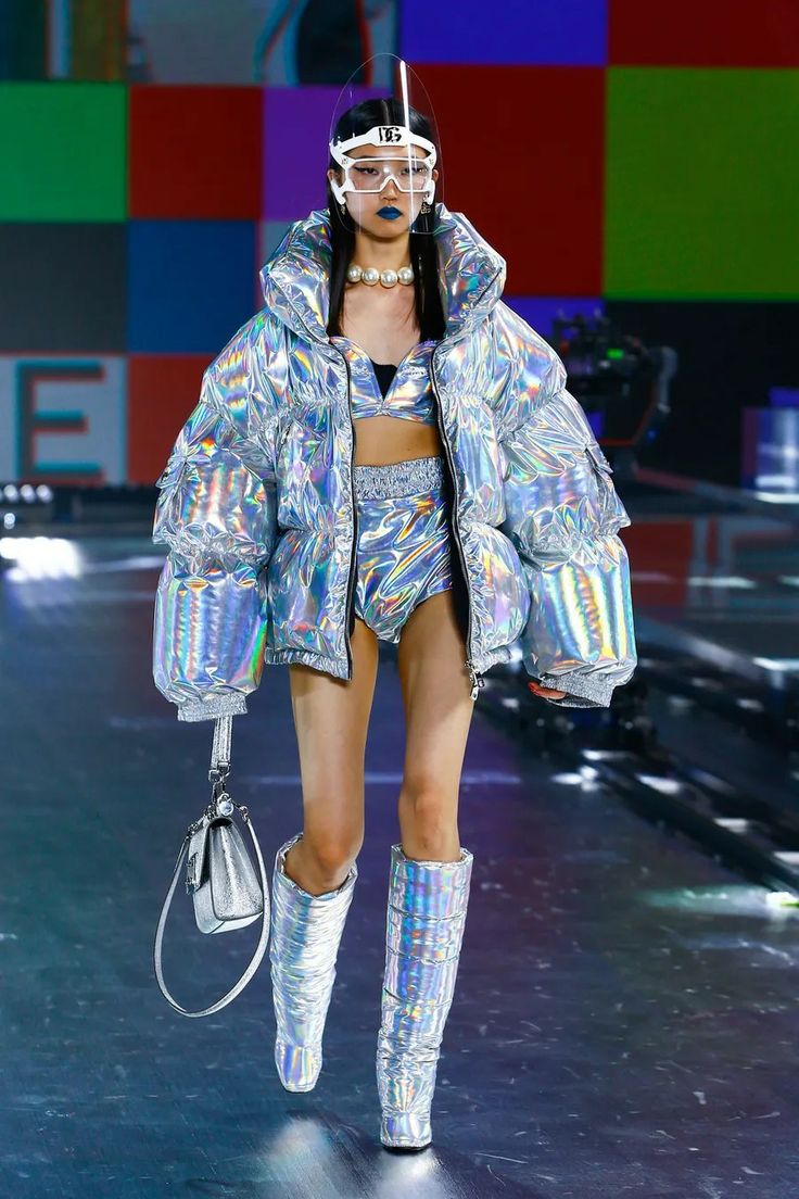 a female model walks down a runway wearing a holographic silver outfit. she has on a puffer jacket, shorts, and boots. she is also wearing a silver handbag and sunglasses. she is wearing blue eye shadow and black eyeliner. the background is a colorful backdrop with large squares.