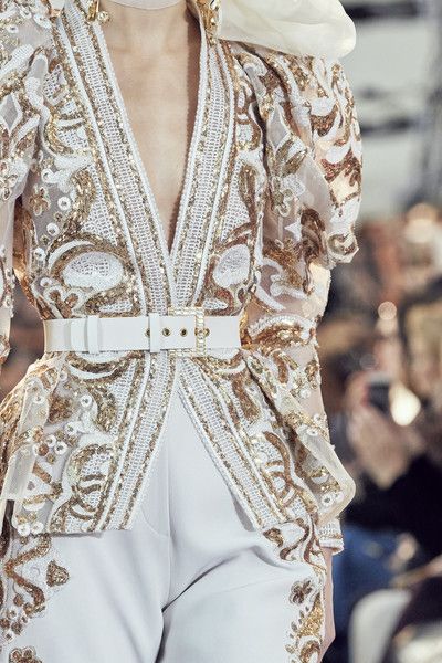 loseup view of a model showcasing a stunning white jumpsuit adorned with intricate gold embroidery and sequins. the jumpsuit features a vneckline, long sleeves with puff detailing at the shoulders, and a fitted waist accentuated by a white belt.  the background is blurred, suggesting a fashion show or runway setting. the overall impression is one of luxurious and elegant high fashion.