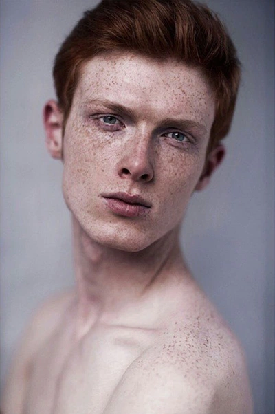 a young, shirtless man with red hair and freckles is captured in a portrait. he has light skin and is looking slightly offcenter with a subtle expression. the focus is on his face and upper body, with the background being a muted, neutral tone.