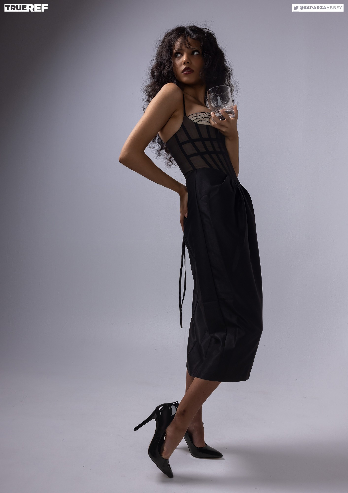 a fulllength studio shot of a woman model, showcasing a black corset dress and high heels against a neutral grey backdrop.  the model is posing, her long dark curly hair cascading over her shoulders, as she confidently holds a glass. the image is stylish, elegant, and captures a confident and sensual mood.  the "true ref" and social media handle are subtly visible.