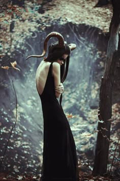 a woman with horns on her head stands in a dark forest, her dark dress flowing around her, in front of a large tree. she has a mysterious and alluring air about her.  she is surrounded by fall leaves, adding to the gothic, mystical, and dramatic atmosphere of the photograph.
