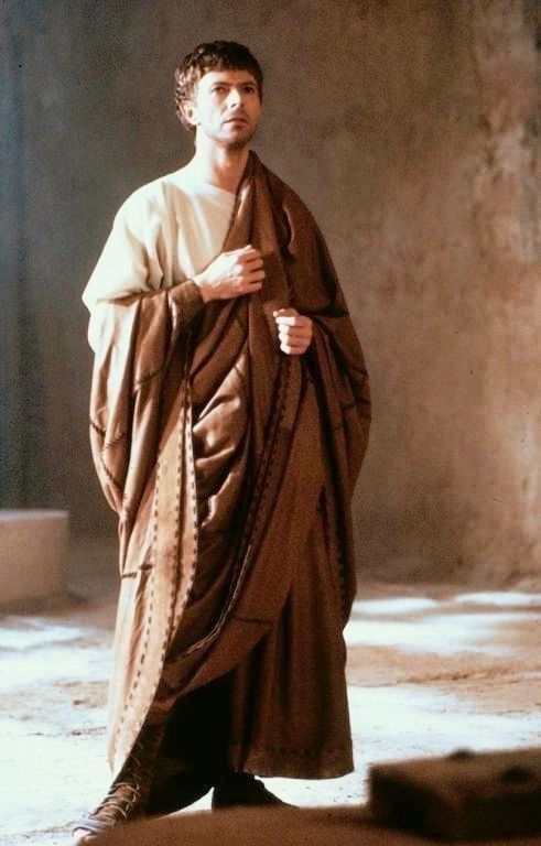 a man dressed in a romanstyle toga stands indoors, wearing a simple white tunic underneath. he has short brown hair and a serious expression. the brown toga is draped over his body and held at the chest. he is wearing simple sandals. the background appears to be a stone or concrete room with natural lighting.